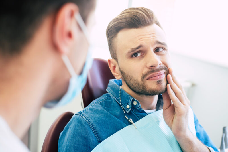Symptoms of gums pain. A man with a worried face is holding his hand on his cheek because of irritating pain in front of a dentist who is going to give a patient a treatment. Man holding his jaw in pain, representing a dental emergency treated at Tampa Palms Dentistry